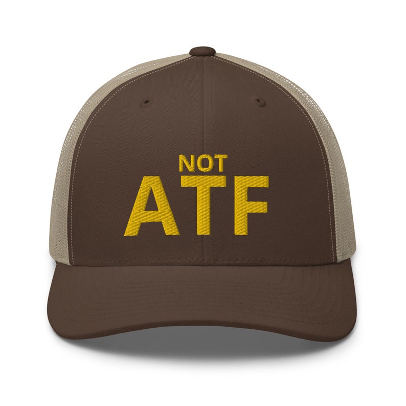 Puede incluir: Una gorra de camionero marr&oacute;n y beige con el texto "NOT ATF" bordado en amarillo.