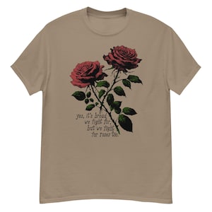 Puede incluir: Una camiseta beige con un gráfico de dos rosas rojas con hojas y tallos verdes. El texto "yes, it's bread we fight for, but we fight for roses too." está impreso debajo de las rosas.