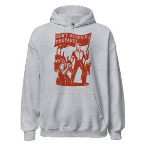 Op de afbeelding: Een lichtgrijze hoodie met een rode afbeelding. De afbeelding toont een persoon die een spandoek vasthoudt met de tekst "DON'T DESPAIR, PREPARE!" en een groep mensen. De hoodie heeft een capuchon en een voorzak.