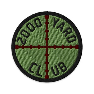 May include: An embroidered patch in green and black with a red crosshair target in the centre. The text "2000 YARD CLUB" is written around the target.