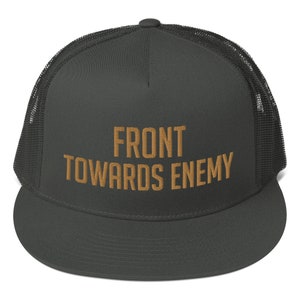 Op de afbeelding: Een donkergrijze truckercap met een zwart mesh-achterpaneel. De pet heeft de tekst "FRONT TOWARDS ENEMY" in goud geborduurd op de voorkant.