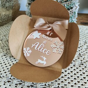 Può includere: Un ornamento natalizio a specchio color oro rosa con un design a fiocco di neve bianco e il testo "Il Natale di Alice". L'ornamento è in una busta di carta marrone.