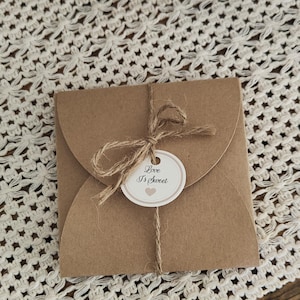 Può includere: Una busta di carta marrone legata con spago, con un piccolo tag circolare che recita "Love Is Sweet" con un cuore. La busta è su una superficie bianca all'uncinetto.