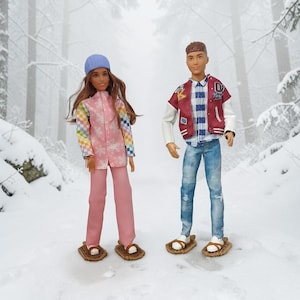 May include: Two dolls in winter clothing stand in a snowy forest. One doll wears a pink snowsuit with snowflake details, a blue hat, and pink trousers. The other doll wears a red and white jacket, blue jeans, and a plaid shirt. Both dolls wear brown and white snowshoes.