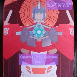 May include: Rectangular mouse pad featuring a stylized illustration of Optimus Prime. The design includes red, blue, and white colors, with the text "Optimus Prime Mouse Pad". The pad measures 9.5" x 7.9".