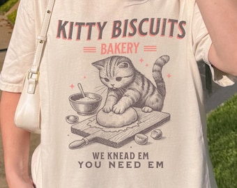 May include: An ivory t-shirt featuring a graphic of a kitten kneading dough on a wooden board. The text reads KITTY BISCUITS BAKERY, WE KNEAD EM YOU NEED EM. The design includes a mixing bowl, a rolling pin, and small stars in brown and red ink.