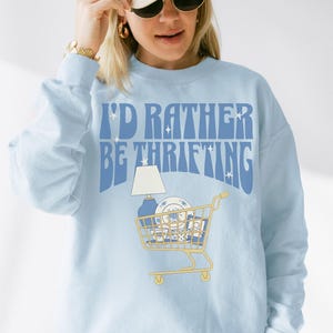 May include: Light blue sweatshirt with the text "I'D RATHER BE THRIFTING" in blue. The graphic features a shopping cart filled with thrifted items, including a lamp and dishes. The sweatshirt is a casual, comfortable style.