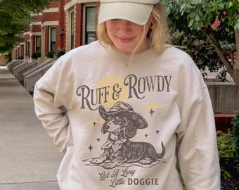 May include: A beige crewneck sweatshirt featuring a graphic of a dachshund wearing a cowboy hat. The text reads "Ruff & Rowdy" and "Get A Long Little DOGGIE" with star accents. The sweatshirt is paired with a matching beige baseball cap.