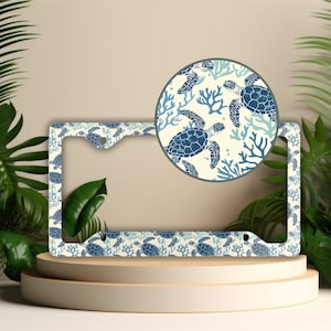 May include: A white license plate frame with a blue and white sea turtle and coral design. The frame is made of metal and has a glossy finish.