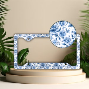 May include: White license plate frame with a blue floral pattern. The frame has two holes on the top and two holes on the bottom for attaching to a license plate.