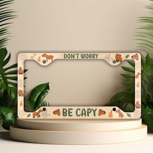 May include: Beige license plate frame with a green border and capybara illustrations. The text "Don't Worry Be Capy" is printed on the frame.