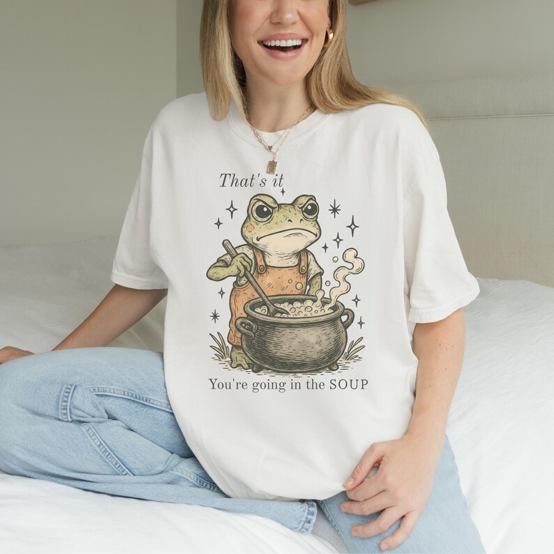 May include: Cream-colored t-shirt featuring a cartoon frog in orange overalls stirring a pot of soup. The text "That's it" and "You're going in the SOUP" are printed above and below the frog illustration. The design includes stars.