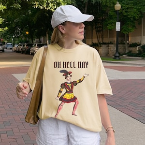 May include: A cream-colored short-sleeve t-shirt featuring a graphic of a historical figure in a colorful doublet and feathered hat, with the text "OH HELL NAY" printed in dark brown lettering above the illustration, paired with white shorts.