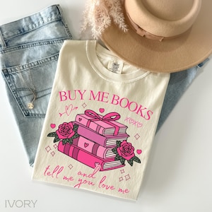 May include: Cream-coloured t-shirt featuring the pink text "BUY ME BOOKS", a stack of books, roses, and hearts. The shirt is styled with light blue jeans and a beige hat.