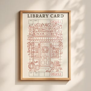 May include: Framed print with a library card design, showcasing a bookstore illustration in brown and beige tones. The print includes the words "LIBRARY CARD" and a bookstore with a wreath and pumpkins. The overall design is autumnal.