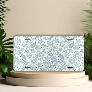 May include: White car registration plate featuring a blue seashell design. The pattern includes a variety of seashells, starfish, and small dots.
