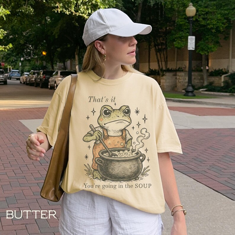 May include: Butter-colored t-shirt featuring a cartoon frog in overalls stirring a pot of soup. Text on the shirt reads "That's it, You're going in the SOUP." The shirt is worn by a person wearing a white cap and shorts.