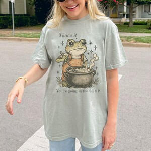 May include: Sage green t-shirt featuring a cartoon frog in overalls stirring a pot of soup. The text "That's it, You're going in the SOUP" is printed above and below the frog illustration. The shirt has a relaxed fit.