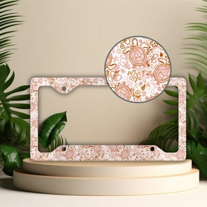 May include: A white license plate frame with a pink and brown floral pattern featuring sea turtles. The frame has two holes for attaching to a license plate.