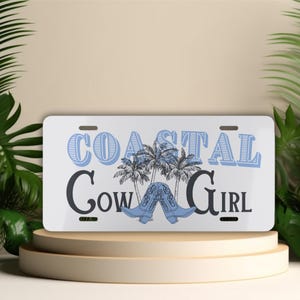 May include: White license plate with the words "COASTAL COW GIRL" in blue and black. The design includes palm trees and blue cowboy boots. The plate is displayed on a beige pedestal.