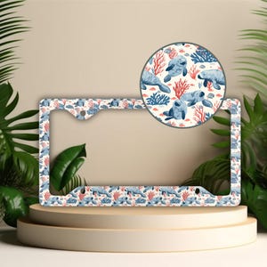 May include: A white metal license plate frame with a colorful pattern of manatees, coral, and fish. The frame is designed to fit a standard US license plate.
