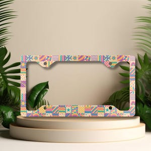 May include: A rectangular license plate frame featuring a colorful patchwork pattern of various geometric shapes, including wavy lines, polka dots, stars, and rainbows in shades of pink, yellow, blue, and orange, with four mounting holes.