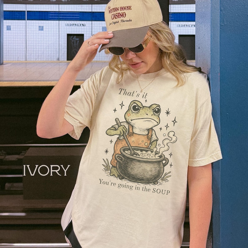 May include: Ivory-colored t-shirt featuring a cartoon frog chef stirring a pot of soup. The text "That's it" is above the frog, and "You're going in the SOUP" is below. The shirt is paired with a beige baseball cap.