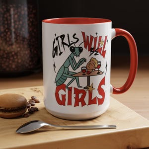 May include: White ceramic mug with a red interior and handle. The mug features a cartoon illustration of a praying mantis and a decapitated head at a table, with the words "GIRLS WILL BE GIRLS" in black and red.