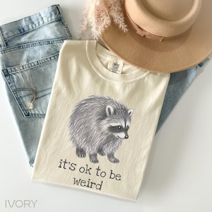 May include: An ivory t-shirt featuring a gray sketch of a raccoon and the black text it's ok to be weird. The shirt has a Comfort Colors label at the inner neckline. The garment is displayed alongside light blue denim jeans and a tan hat.