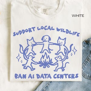 May include: A white t-shirt featuring a blue graphic of a fox, dog, cat, bird, and mouse holding hands in a circle around a campfire. The text reads SUPPORT LOCAL WILDLIFE at the top and BAN AI DATA CENTERS at the bottom in blue lettering.