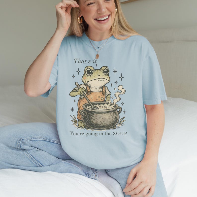 May include: Light blue t-shirt featuring a cartoon frog in orange overalls stirring a pot of soup. The text "That's it" and "You're going in the SOUP" are printed on the shirt. The shirt is made of a soft material.