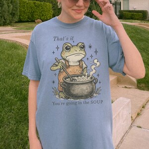 May include: A light blue t-shirt featuring a cartoon frog wearing an apron and stirring a pot of soup. The text "That's it" and "You're going in the SOUP" are printed above and below the frog illustration.