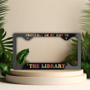 May include: Black license plate frame with the text "Probably on my way to the library" in colorful letters.