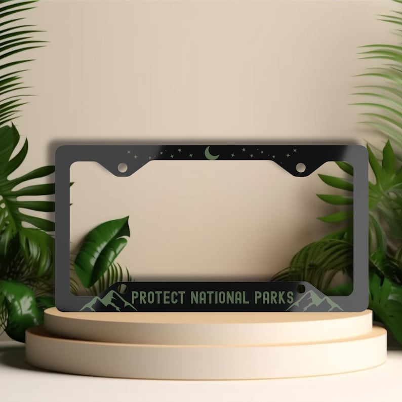 May include: A dark grey license plate frame with a night sky design featuring stars and a crescent moon. The bottom of the frame has a mountain range graphic and the text "PROTECT NATIONAL PARKS".