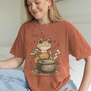 May include: Burnt orange t-shirt featuring a cartoon frog stirring a pot of soup. The frog wears overalls and has an angry expression. Text on the shirt reads "That's it" and "You're going in the SOUP".