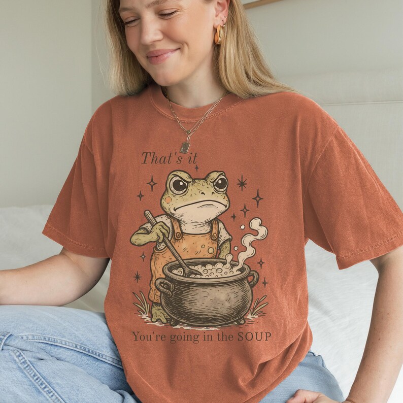 May include: Burnt orange t-shirt featuring a cartoon frog stirring a pot of soup. The frog wears overalls and has an angry expression. Text on the shirt reads "That's it" and "You're going in the SOUP".