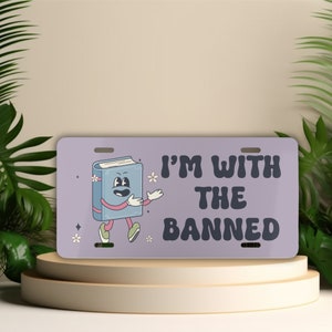Może przedstawiać: Prostokątna tablica rejestracyjna z jasnofioletowym tłem. Zawiera postać z kresk&oacute;wki w postaci książki z rękami i nogami oraz napis "I'M WITH THE BANNED" w kolorze ciemnofioletowym. Małe białe kwiaty i gwiazdki zdobią tablicę.