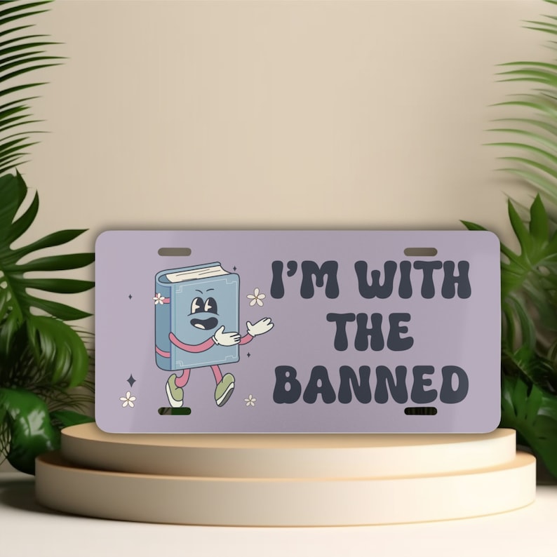 Może przedstawiać: Prostokątna tablica rejestracyjna z jasnofioletowym tłem. Zawiera postać z kresk&oacute;wki w postaci książki z rękami i nogami oraz napis "I'M WITH THE BANNED" w kolorze ciemnofioletowym. Małe białe kwiaty i gwiazdki zdobią tablicę.