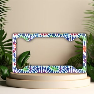 May include: A white license plate frame with a colorful tie-dye pattern. The frame has holes for screws and a metal finish.
