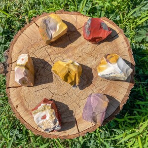 Może przedstawiać: Sześć surowych, nieoszlifowanych kamieni mineralnych ułożonych w okrąg na okrągłym plastrze drewna. Kamienie mają zróżnicowane kolory, w tym musztardowy żółty, głęboki czerwony, biały i delikatny fiolet, z nieregularnymi krawędziami i gładkimi powierzchniami.