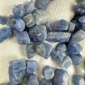Puede incluir: Una colección de piedras preciosas de zafiro azul en bruto. Los zafiros, de varios tamaños y formas, exhiben una gama de tonos azules, desde claros hasta índigo intenso. Las piedras preciosas están dispuestas sobre una superficie blanca texturizada, mostrando su estado natural sin pulir.