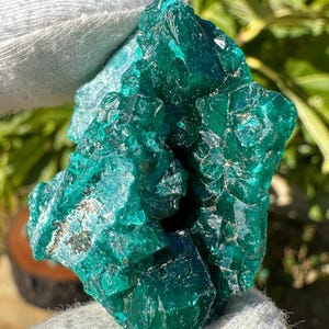 Könnte beinhalten: Ein rohes, leuchtend blaugrünes Mineralexemplar, bestehend aus einer Ansammlung kantiger, durchscheinender Kristalle mit glasartigem Glanz. Der Stein zeigt tiefe smaragdgrüne Farbtöne und scharfe, geometrische Facetten in seiner komplexen Struktur.