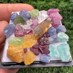 Peut inclure: Une collection de pierres précieuses brutes et multicolores disposées sur un support blanc. L'assortiment comprend des cristaux de formes et de tailles variées en bleu, jaune, vert, rose et transparent, ainsi qu'un cristal brun translucide allongé.