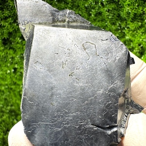 Könnte beinhalten: Dunkelgraues, unregelmäßig geformtes Mineralexemplar mit strukturierter Oberfläche und scharfen Kanten. Der Stein hat ein facettiertes Aussehen und eine leicht reflektierende Qualität. Der Hintergrund ist ein verschwommenes Grün.