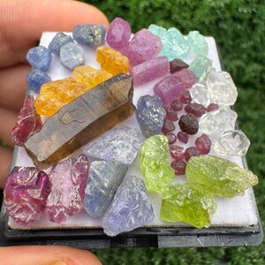 May include: A collection of assorted raw, uncut gemstones in various colours including yellow, blue, pink, green, and clear, arranged on a white square display pad. The assortment features a prominent dark brown rectangular crystal piece.