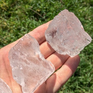 May include: Two clear quartz crystals, one larger than the other, are held in a hand against a green grassy background.