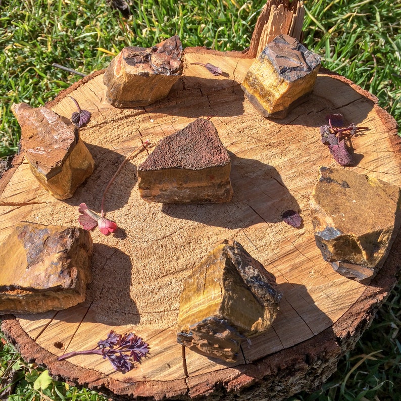 May include: Six tiger's eye stones of various sizes and shapes arranged on a wooden surface. The stones are a mix of brown, gold, and black.