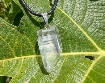 Puede incluir: Un colgante de cristal transparente con una fianza plateada cuelga de un collar de cordón negro. El cristal está facetado y tiene un aspecto natural y en bruto. El colgante se muestra sobre un fondo de hojas verdes, destacando su claridad y forma.