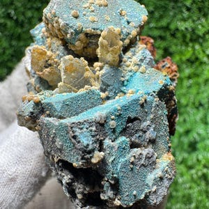 May include: A close-up of a mineral specimen featuring a mix of colors and textures. The rock has a base of dark gray and black, with patches of vibrant turquoise and areas of tan and clear crystal formations.