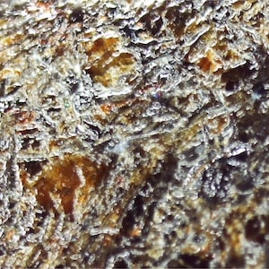 May include: A close-up view of a rough, textured mineral surface featuring a complex network of deep crevices and raised ridges. The material displays a variegated colour palette of dark brown, amber, and metallic grey tones with a crystalline sheen.